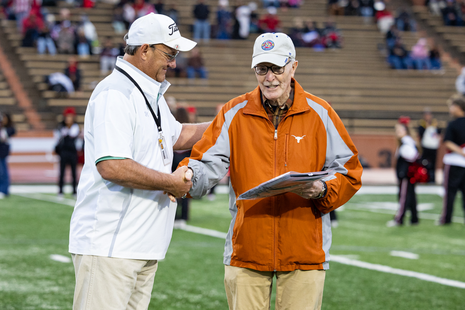 THS-AHS-FB-1 | Press Pass Sports Two men shake hands on a football field; one in a white jacket and cap, the other in an orange-gray jacket holding a clipboard.