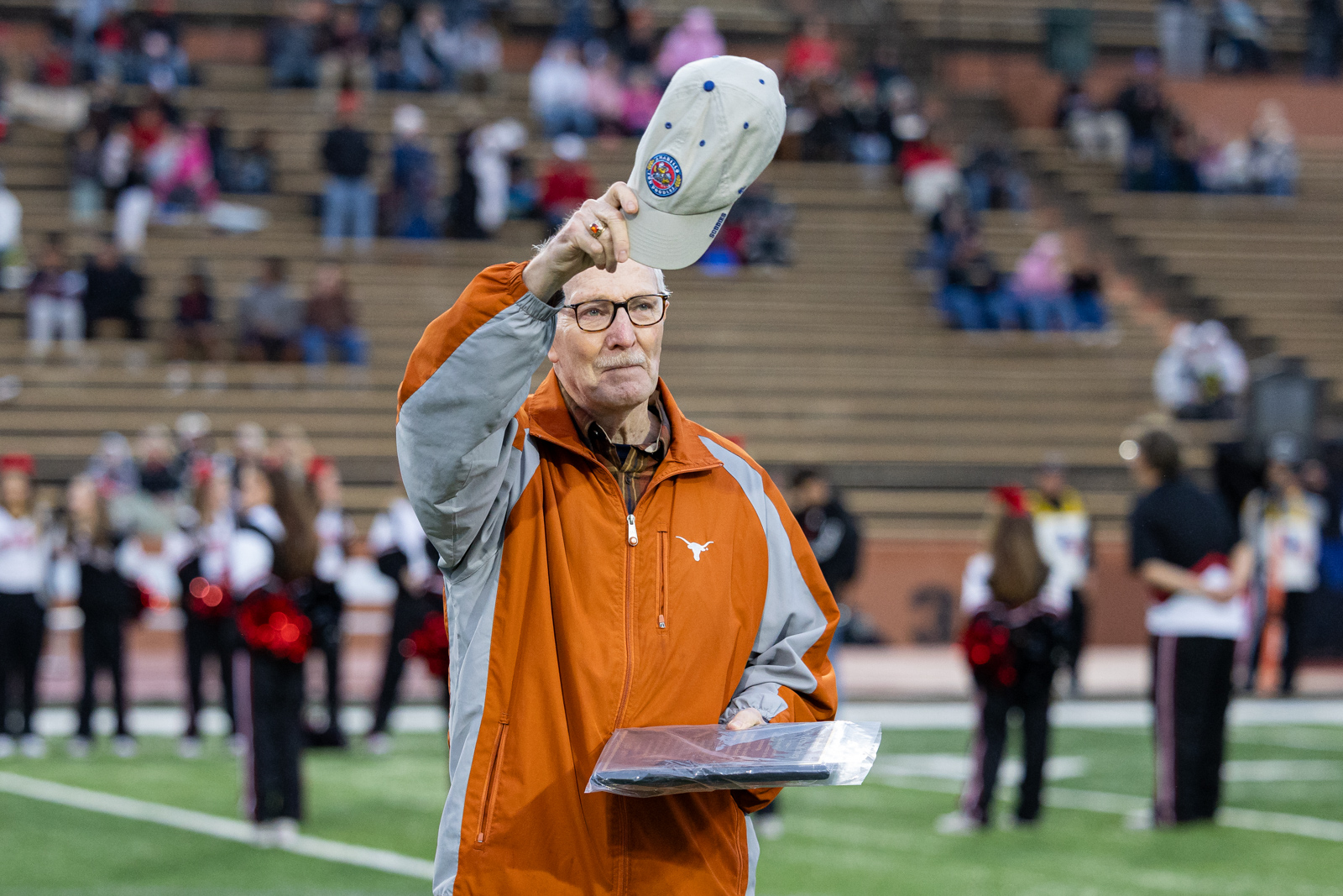 THS-AHS-FB-2 | Press Pass Sports Older man in an orange and gray jacket raises a light-colored cap into the air on a football field, holding a clipboard or folder.