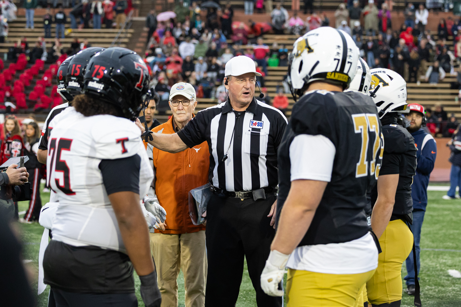 THS-AHS-FB-4 | Press Pass Sports Referee in black-and-white stripes speaks to football players on the field as teammates listen, with a crowd in the stands behind.