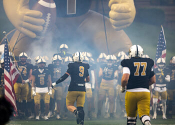 THS-AHS-FB-5 | Press Pass Sports Football team enters the field through blue-gray smoke, leading with #9 ahead of teammates in black and gold uniforms, inflatable mascot in the background