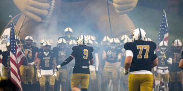 Football team enters the field through blue-gray smoke, leading with #9 ahead of teammates in black and gold uniforms, inflatable mascot in the background