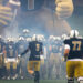 THS-AHS-FB-5 | Press Pass Sports Football team enters the field through blue-gray smoke, leading with #9 ahead of teammates in black and gold uniforms, inflatable mascot in the background