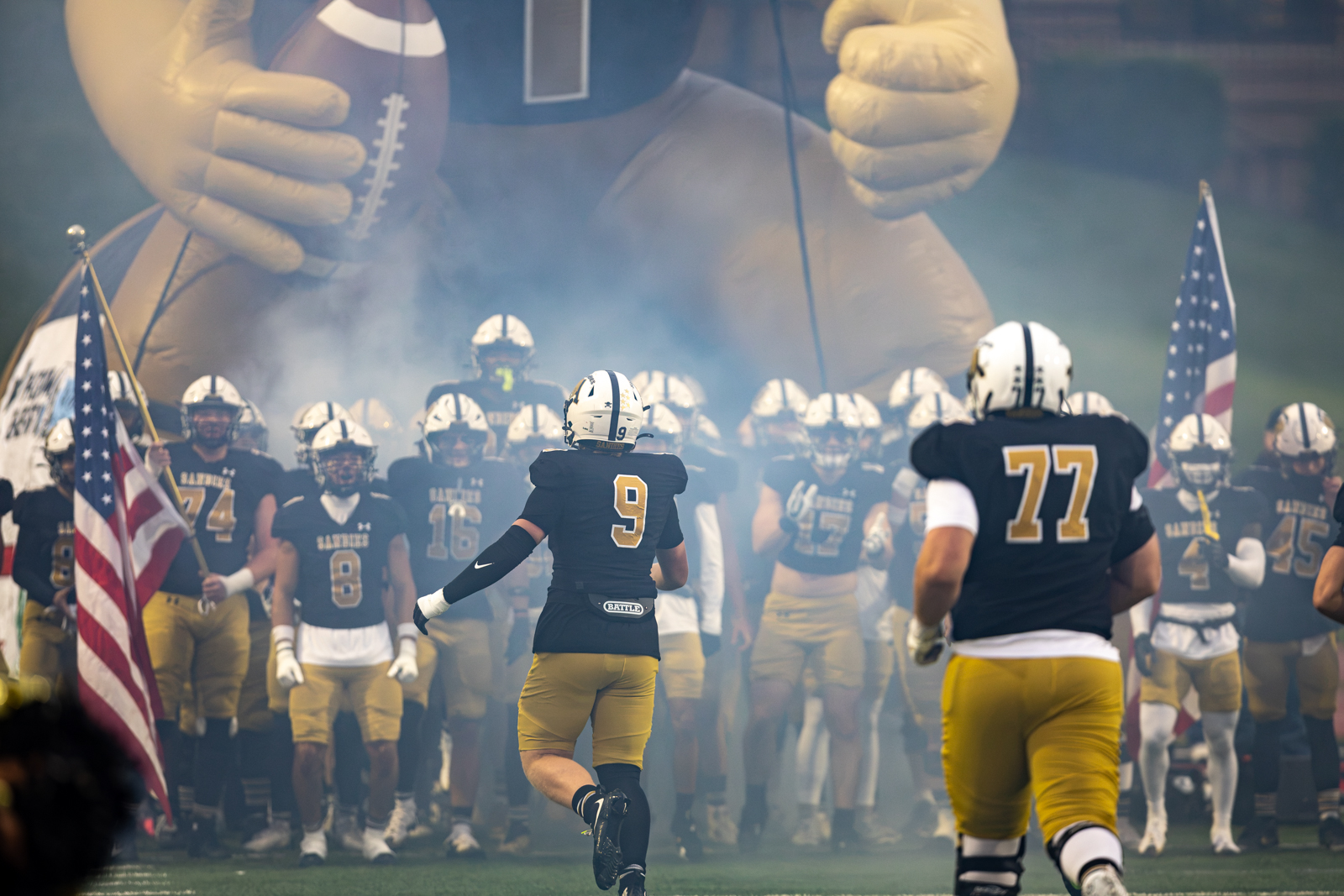 THS-AHS-FB-5 | Press Pass Sports Football team enters the field through blue-gray smoke, leading with #9 ahead of teammates in black and gold uniforms, inflatable mascot in the background