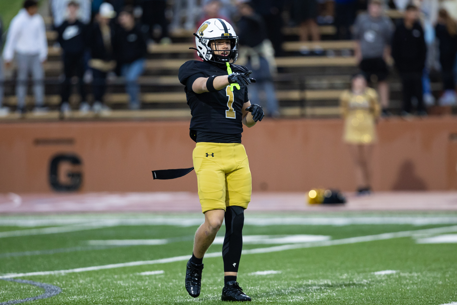 THS-AHS-FB-6 | Press Pass Sports Football player in a black jersey and yellow shorts points during a game on a green field with spectators in the stands behind him.