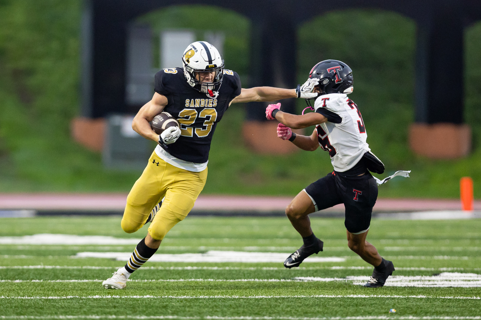 THS-AHS-FB-7 | Press Pass Sports A football running back in black and gold carries the ball while a defender in white and red reaches in for a tackle on a green field.