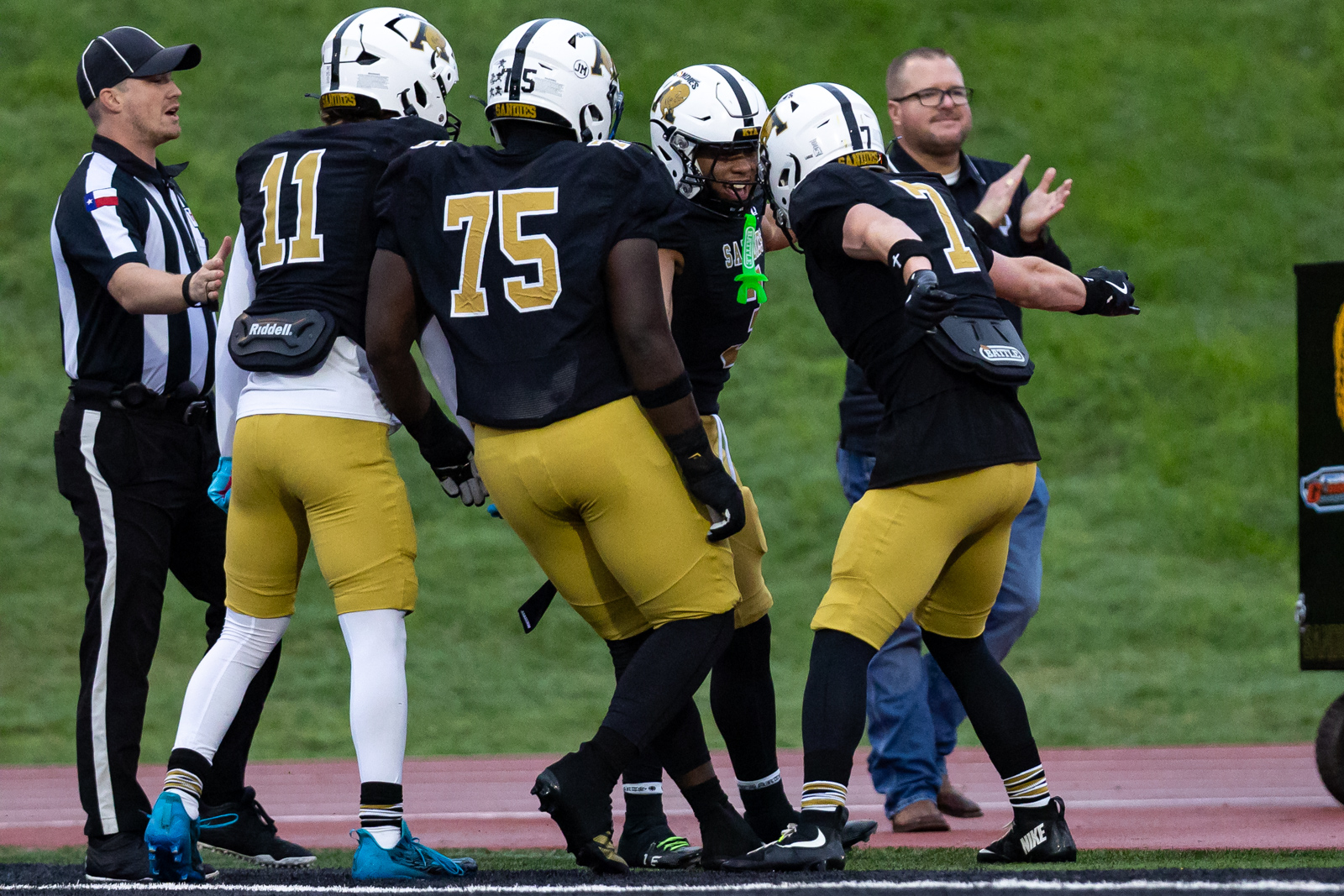 THS-AHS-FB-10 | Press Pass Sports American football players in black and gold uniforms huddle on a track, referee nearby, celebrating a play on the field