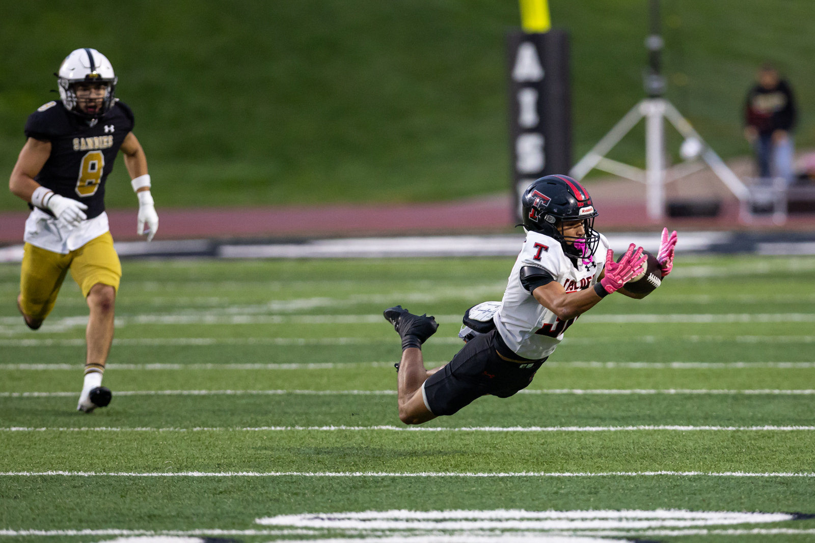 THS-AHS-FB-12 | Press Pass Sports Wide receiver in white uniform dives for a catch, pink gloves visible, while a defender in black and gold chases on a green field behind him.