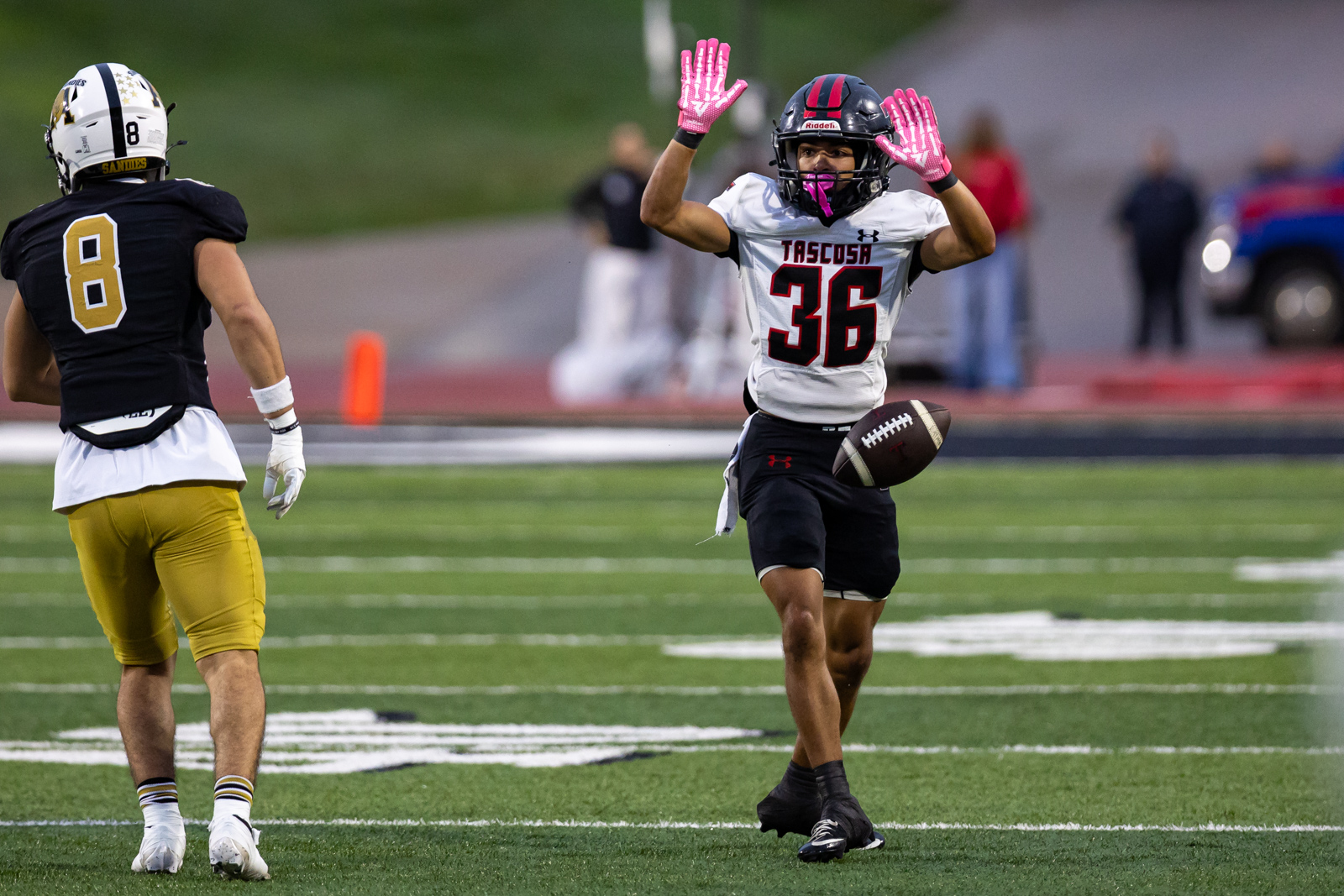 THS-AHS-FB-13 | Press Pass Sports Football game action: Tasosa #36 in white jersey with pink gloves raises hands as he prepares to advance, holding the football; defender in black and gold #8 faces away.