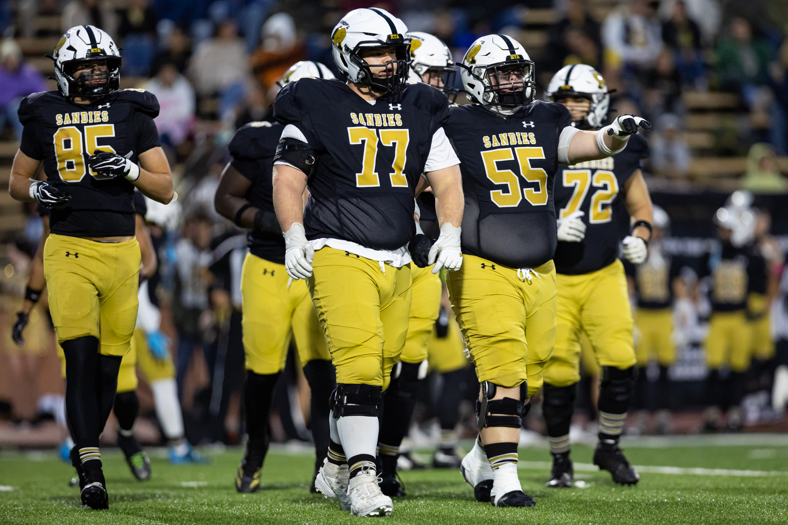 THS-AHS-FB-17 | Press Pass Sports Sandies football players in black jerseys and yellow pants walk onto a green field during a game.