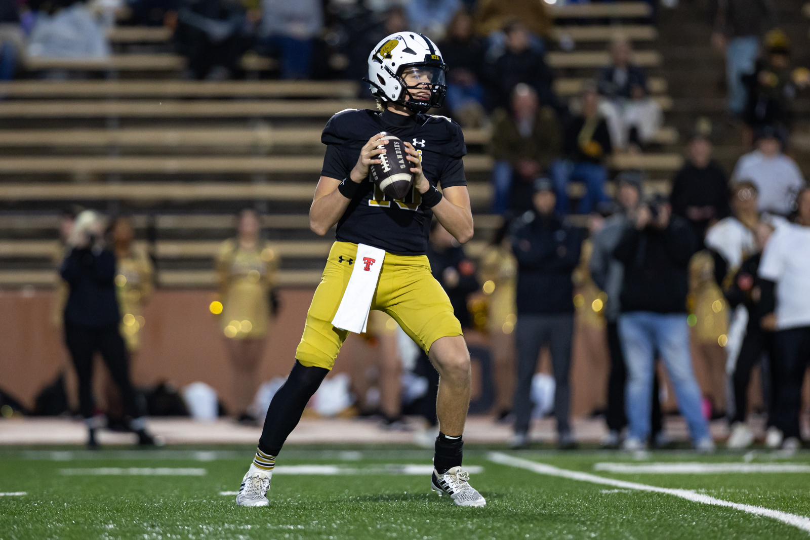 THS-AHS-FB-20 | Press Pass Sports Quarterback in a black jersey and gold pants drops back to pass, gripping a football on a crowded sideline field.
