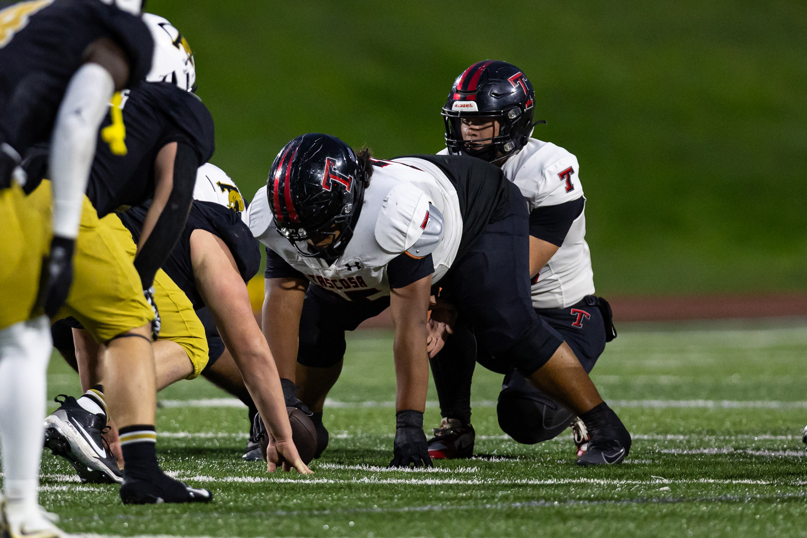 THS-AHS-FB-23 | Press Pass Sports American football players line up at the snap; the quarterback center bends over the football behind him, teammates ready to block or rush.