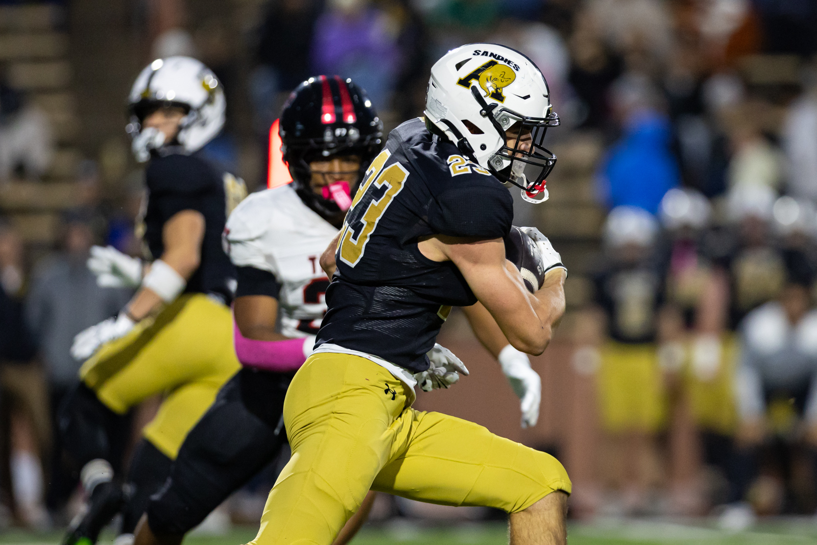 THS-AHS-FB-24 | Press Pass Sports Football player in a black jersey and gold pants runs with the ball, white helmet with a yellow mascot, #23, during a game with defenders in pursuit nearby