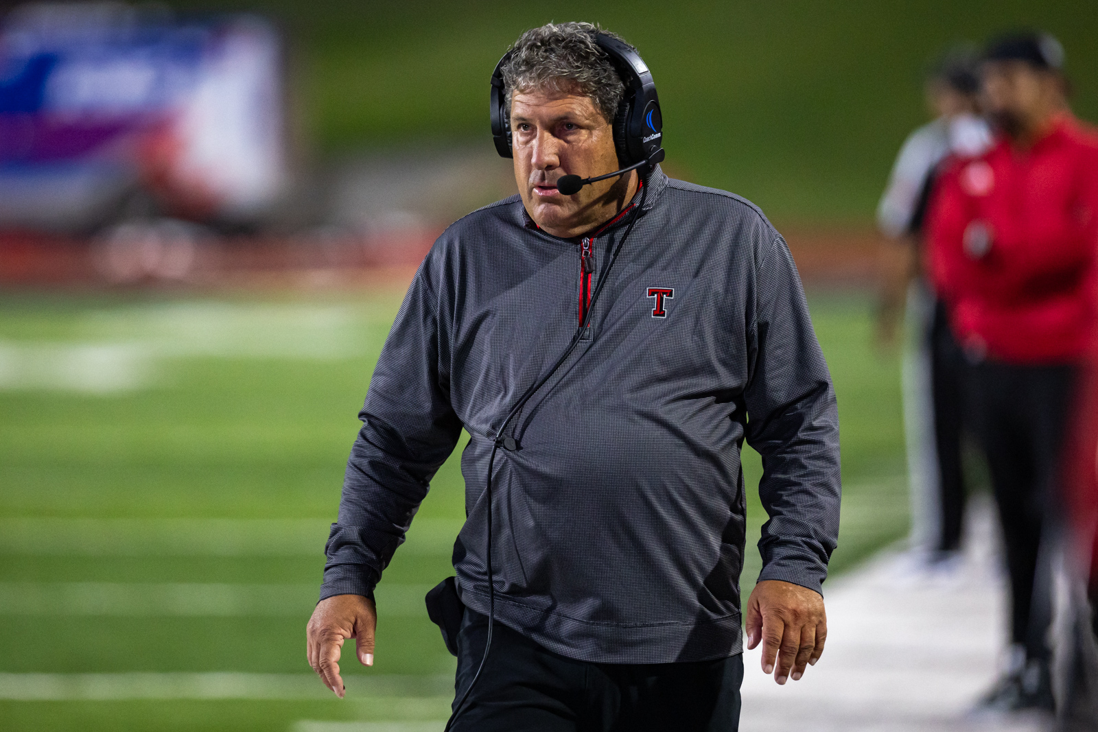THS-AHS-FB-25 | Press Pass Sports Football coach wearing a headset and gray pullover walks along the sideline during a game, focused and alert.