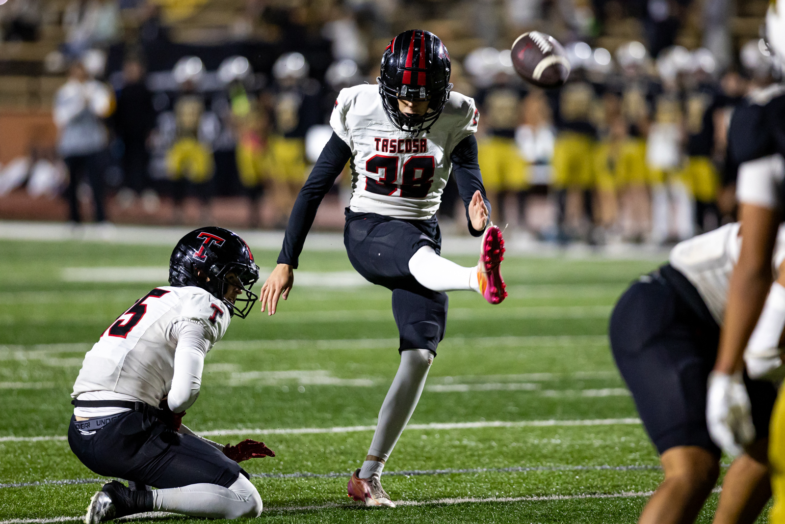 THS-AHS-FB-29 | Press Pass Sports A football kicker in white jersey numbered 39 leaps to kick the ball as a tackleer in white jersey 53 dives nearby on a green turf field at night. This conveys action: kicking play during a game.