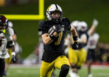 THS-AHS-FB-30 | Press Pass Sports Football player wearing a black jersey (number 7) and yellow pants runs with the ball during a game at night stadium.
