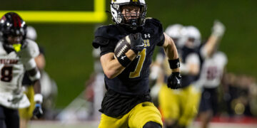 Football player wearing a black jersey (number 7) and yellow pants runs with the ball during a game at night stadium.