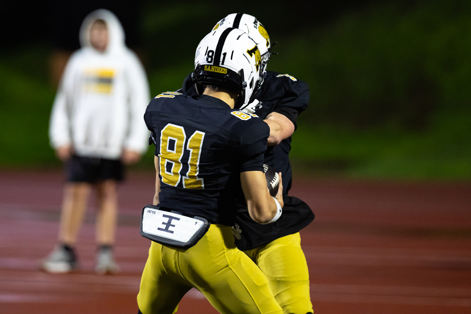 THS-AHS-FB-32 | Press Pass Sports Two football players in black and gold collide during a night game on a track, with the number 81 visible on one jersey.