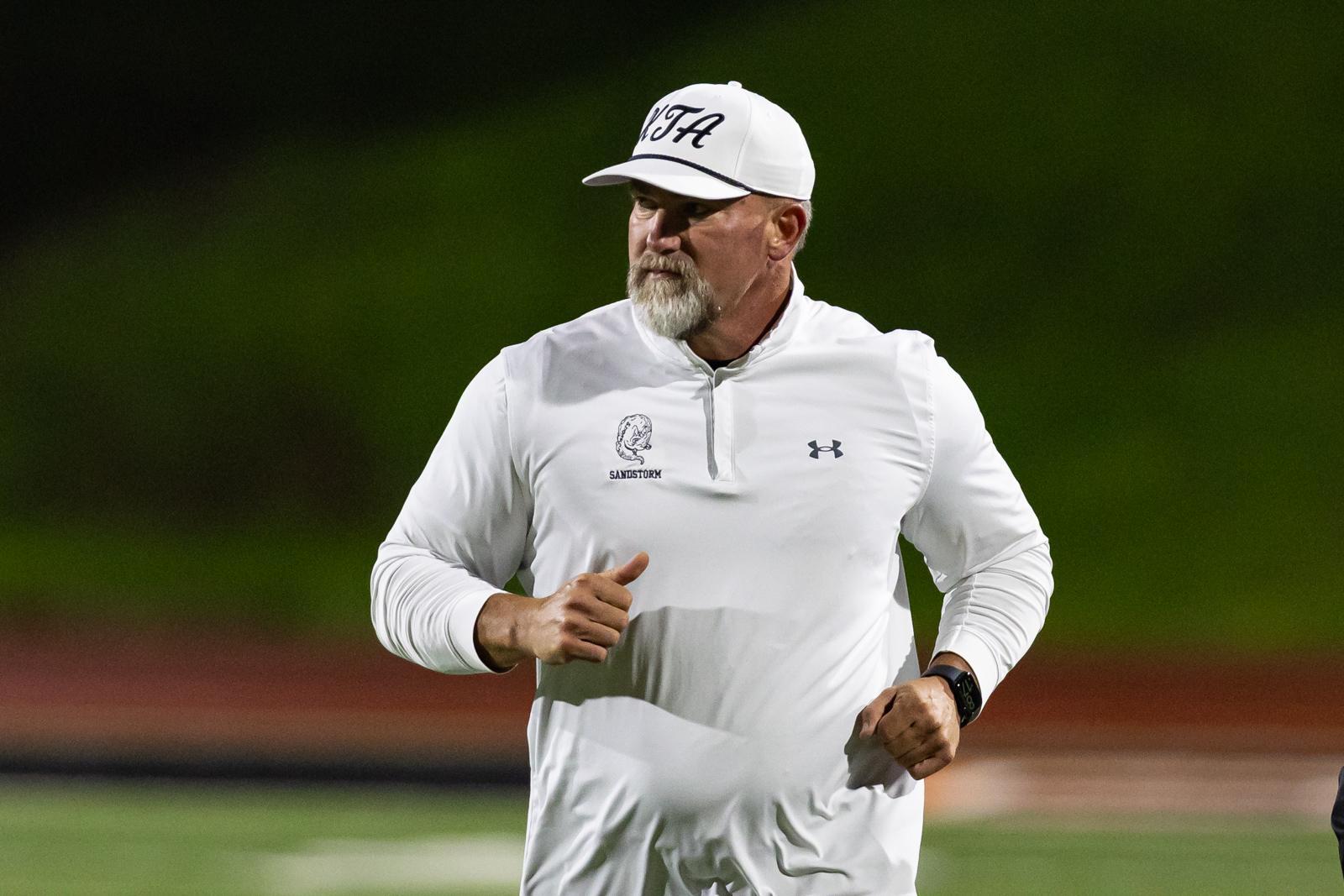 THS-AHS-FB-40 | Press Pass Sports Man in a white athletic top and cap jogging on a sports field at night, left arm bent and focused expression on his face.
