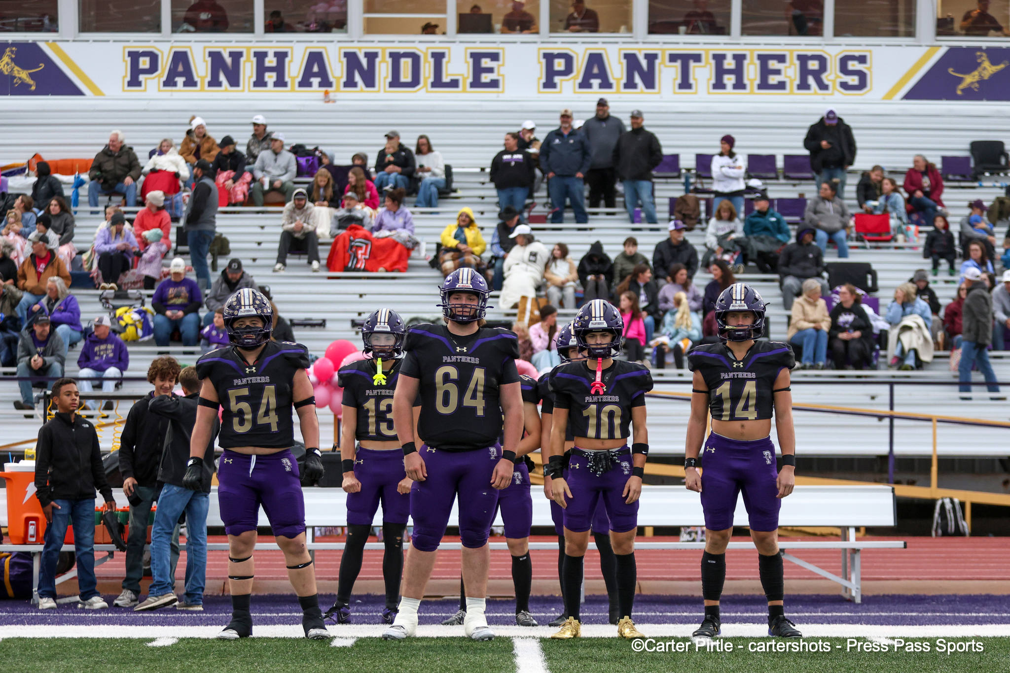 High school football players in purple uniforms stand in a line on the field with bleachers full of spectators behind them.