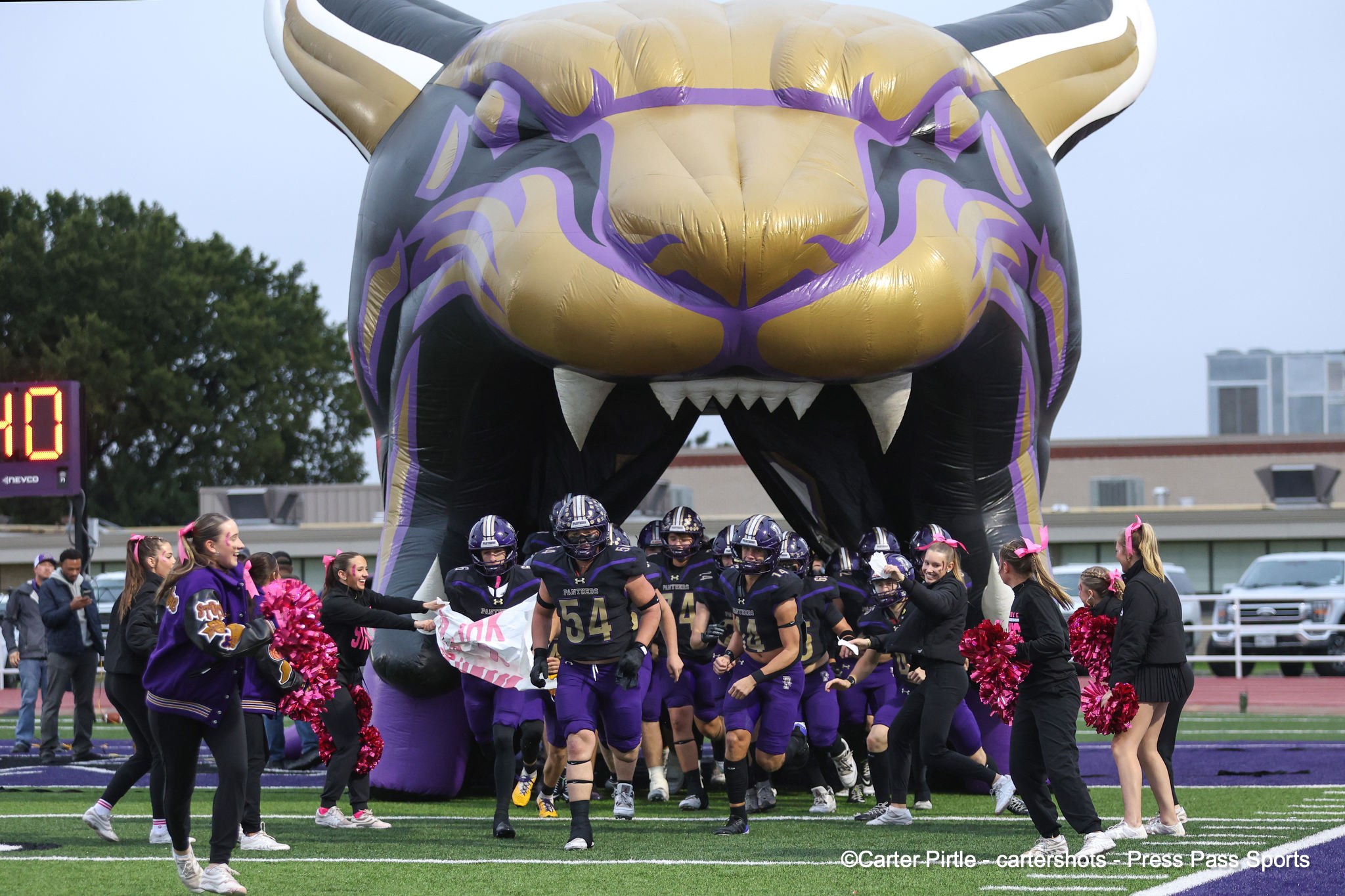 Football players in purple uniforms sprint onto the field from a giant gold and purple inflatable tiger head, with cheerleaders nearby.