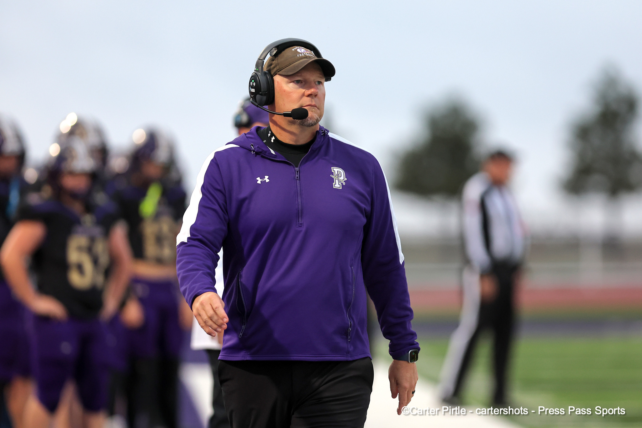 Coach wearing a headset in a purple jacket on a football field sidelines