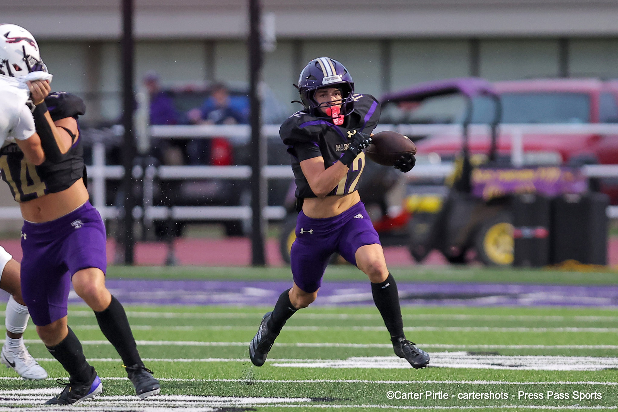 Female football player in purple and black runs with the ball during a game on a turf field, others visible in the background.