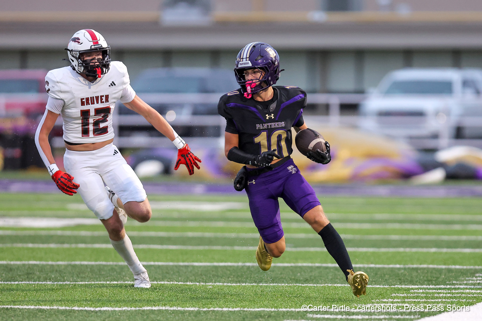 Two football players sprint on a grassy field during a game: left player in white with number 12, right player in purple Panthers with number 10 holding the ball.