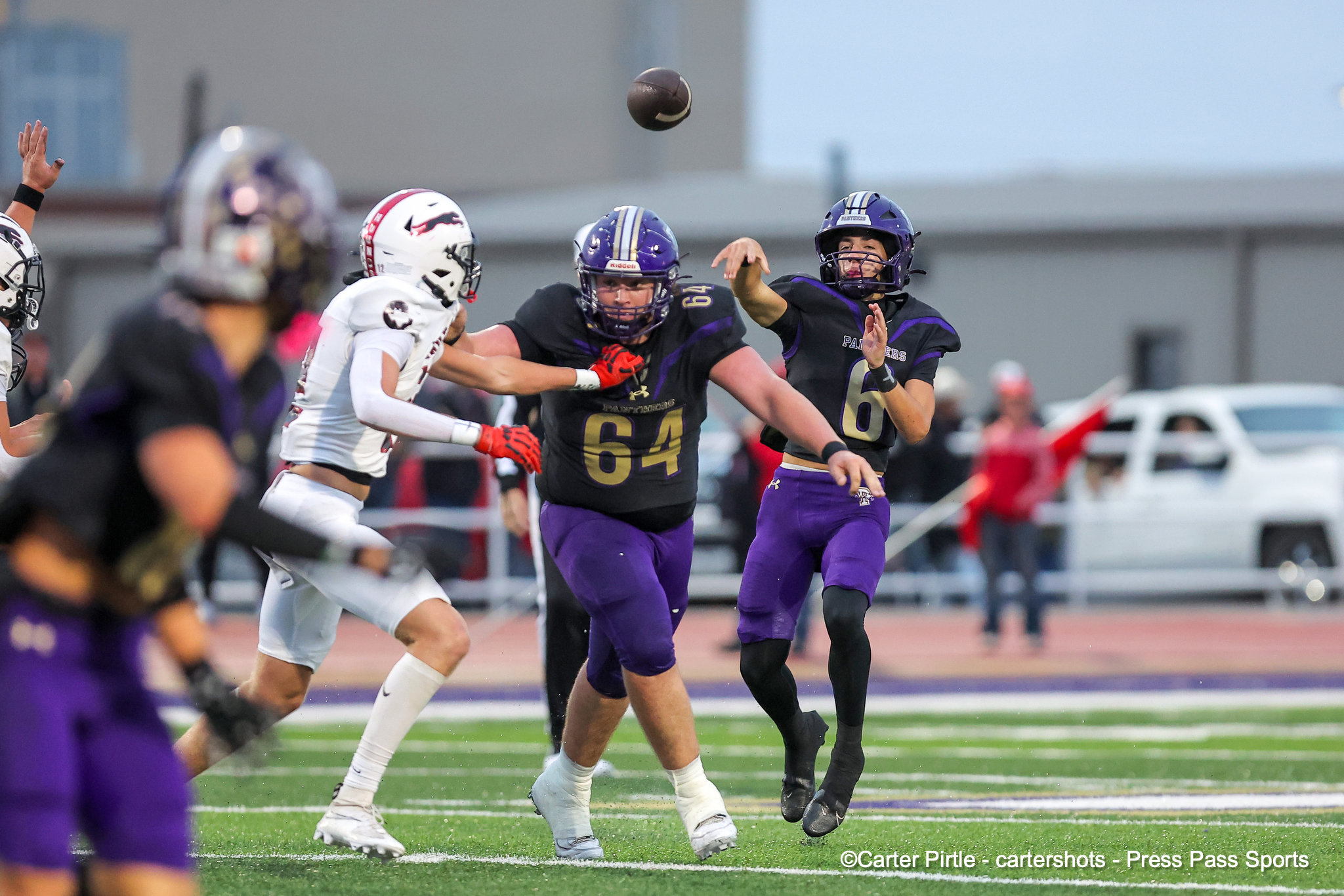 Two football players in purple uniforms reach for a high pass as the black ball sails between them on a green turf field, with white-uniform defenders nearby.