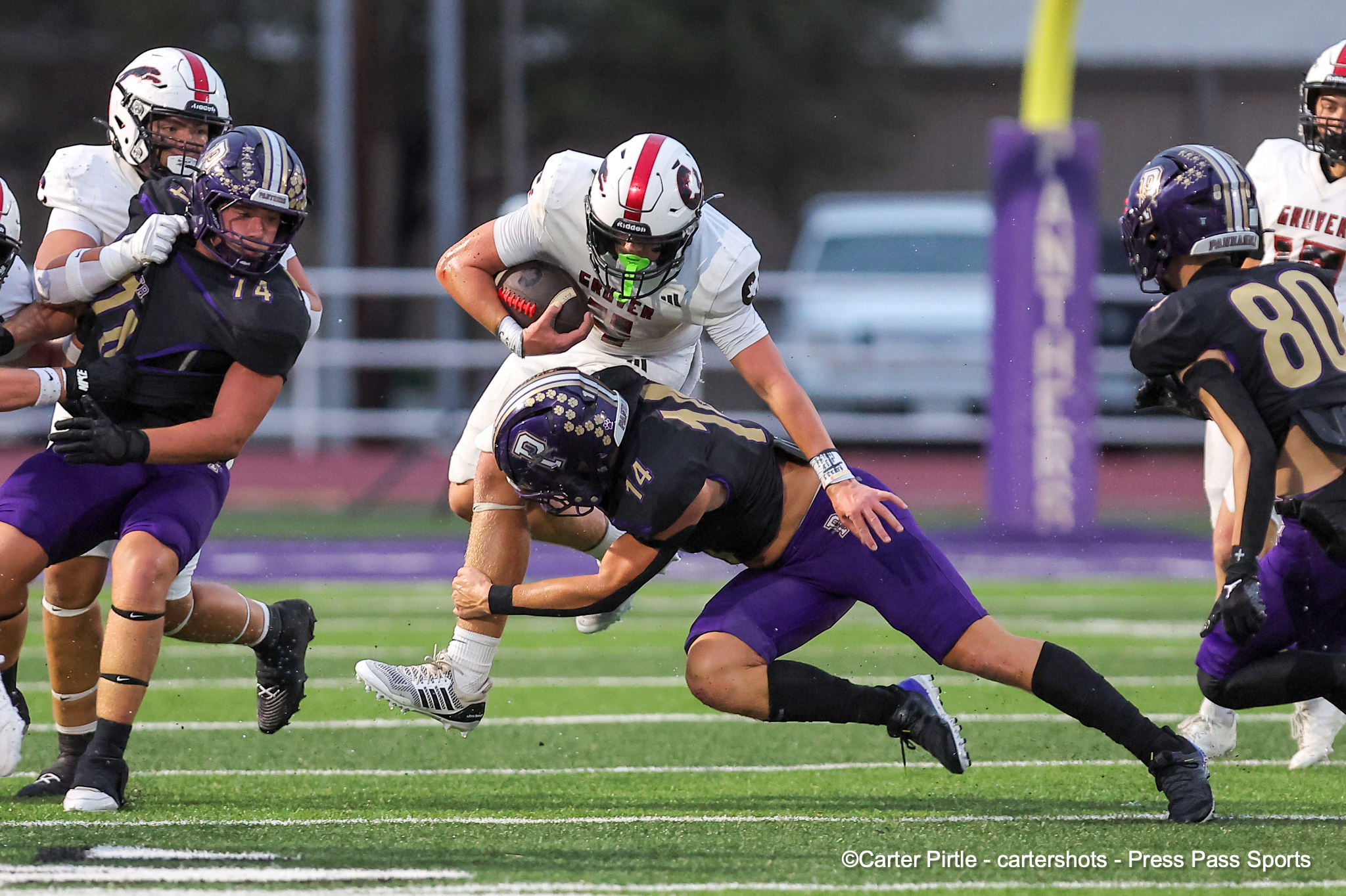 Football players in white and purple collide as a ball carrier in white runs forward while a purple defender lunges in for the tackle on a green field.