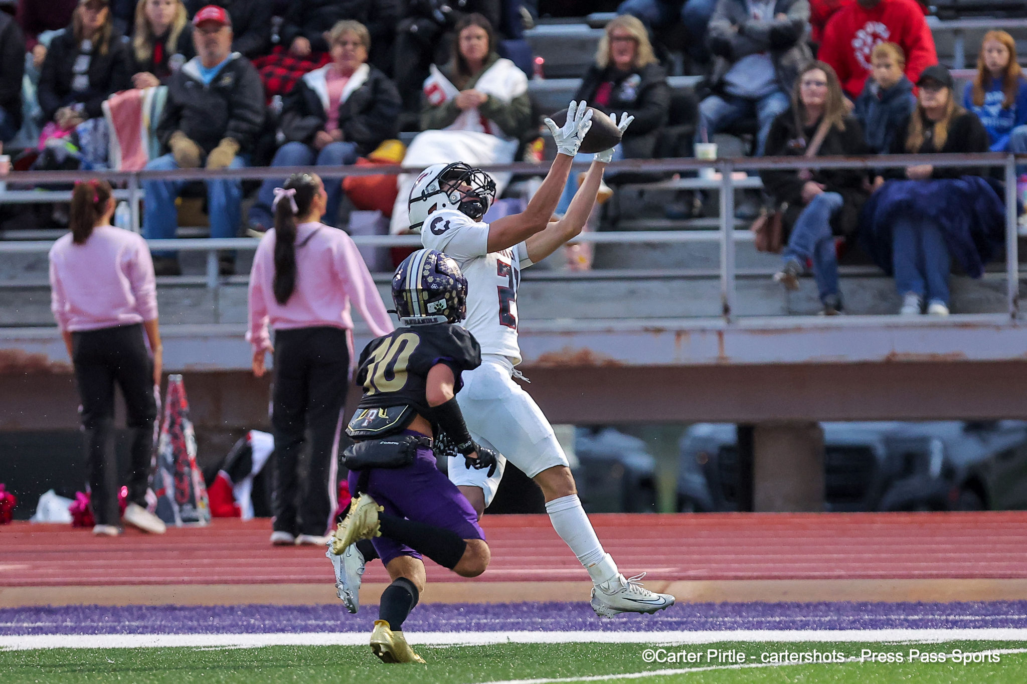 American football action: a white-jersey player reaches up to catch or intercept the ball while a defender in black and purple pursues, on a track field with spectators in the bleachers behind.