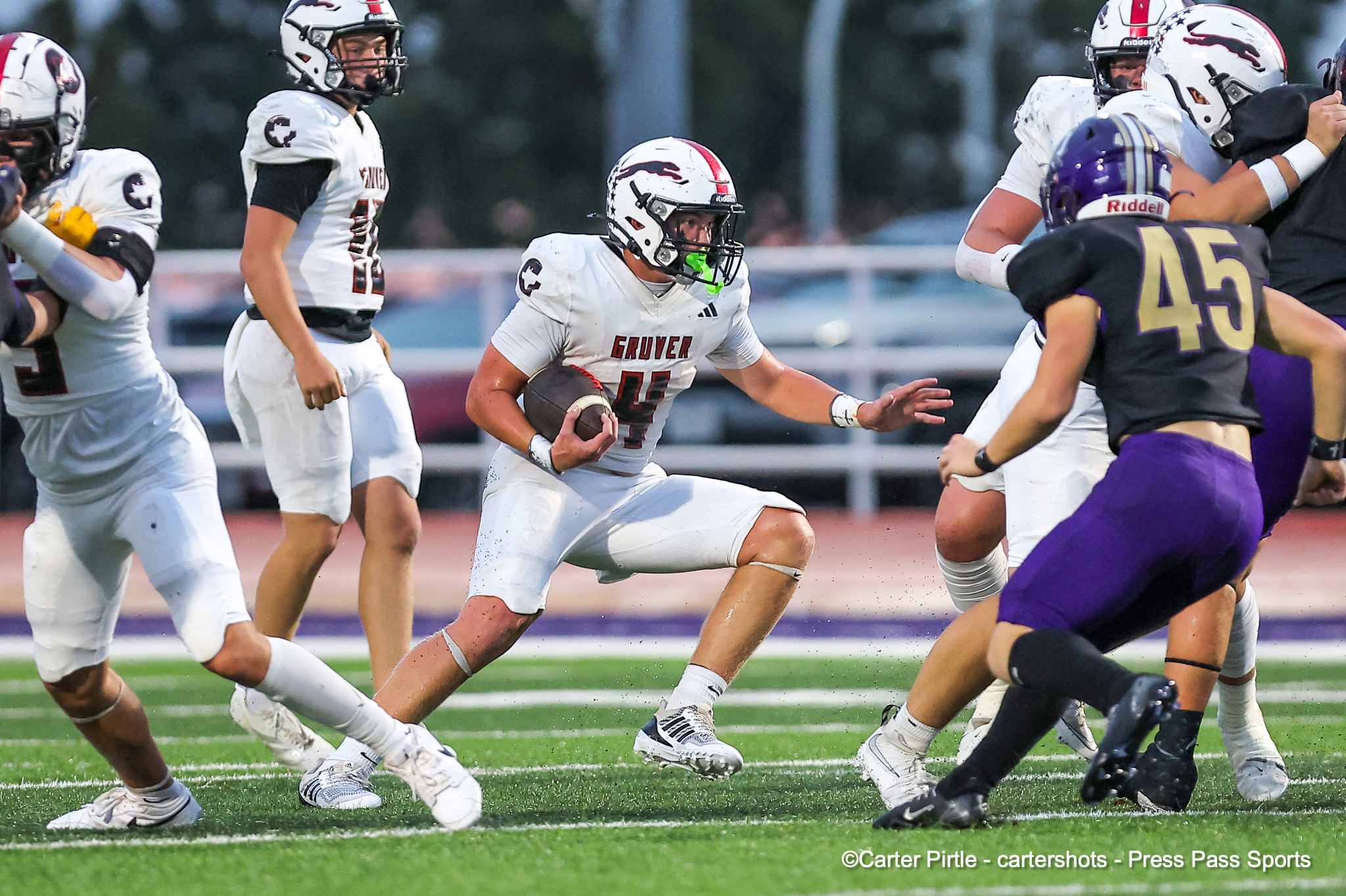 Football game action: white-team ball carrier runs with the ball while defenders in purple close in on him on a green field.