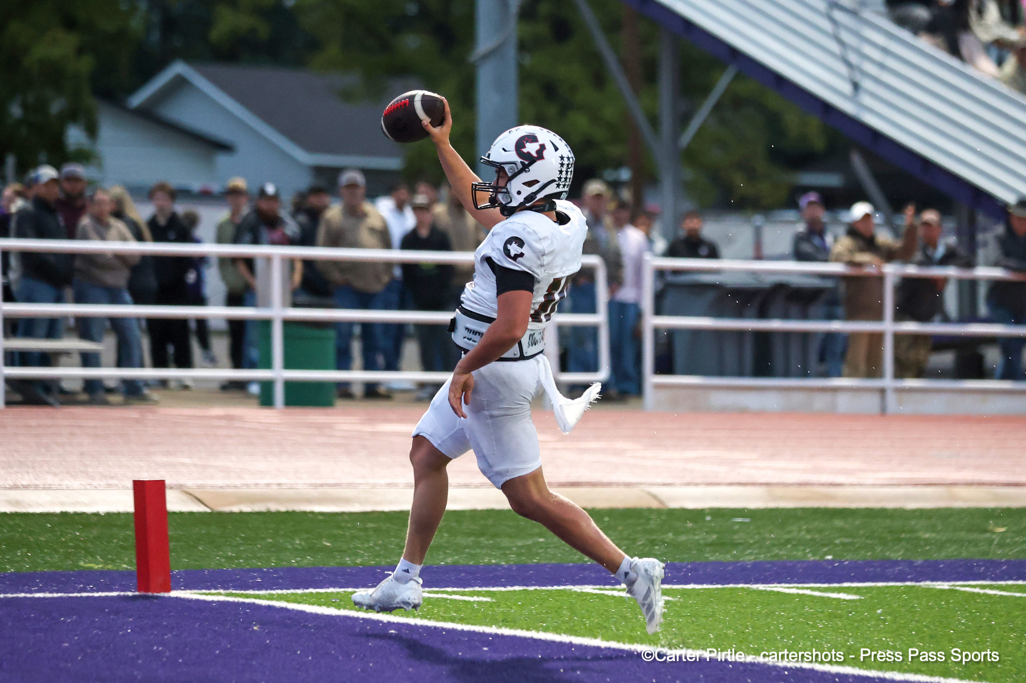 Football player in white uniform lifts the ball to throw on a purple-turf field, with spectators behind a white fence.