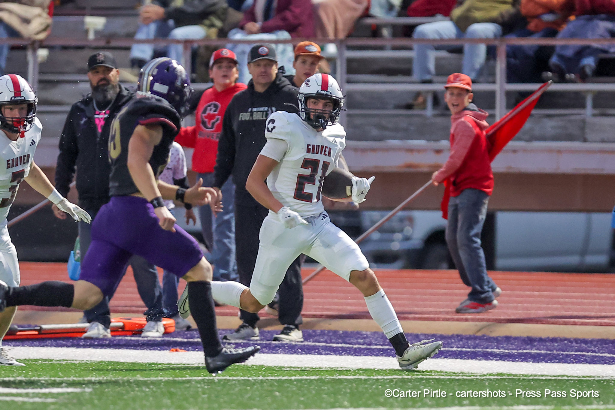 White-jersey football player #21 runs with the ball as teammates and defenders chase on a purple turf field, crowd in the stands.