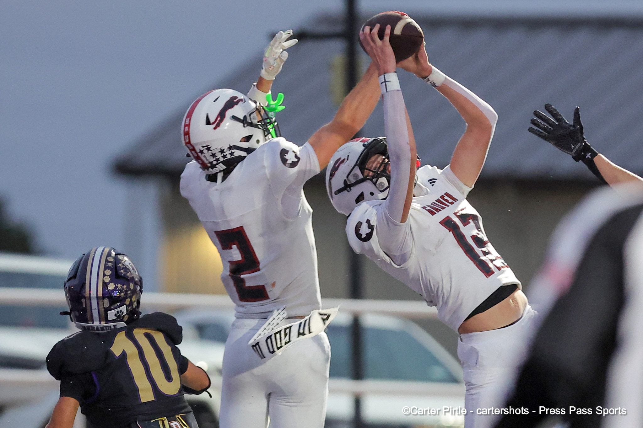 Two football players in white jerseys leap and reach for a airborne ball during a play, with a defender in black nearby while a purple-helmeted player watches.