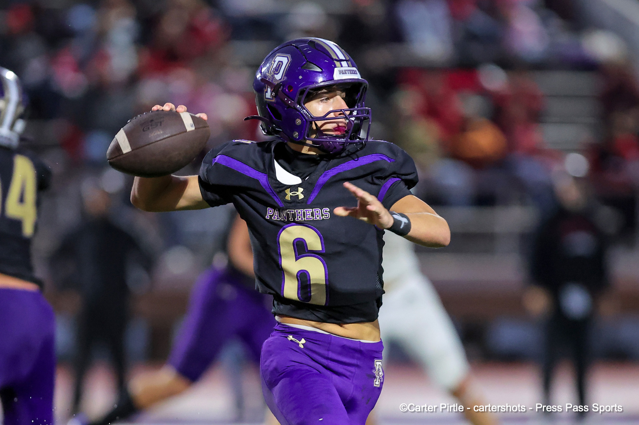 Football quarterback in a purple uniform (number 6) throwing a pass during a game in a crowded stadium with teammates in purple nearby and a purple helmet.