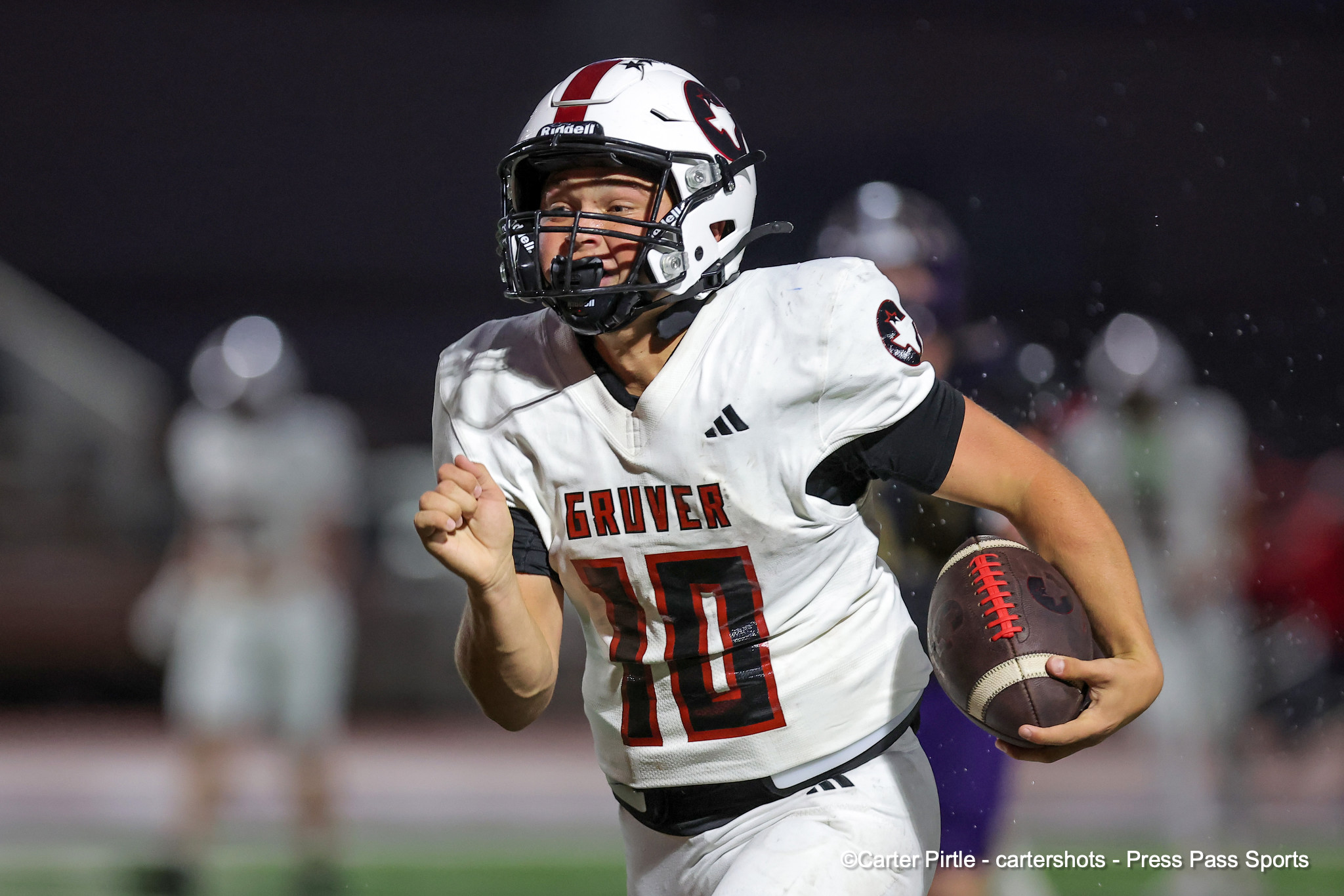 Young football player in a white uniform with red lettering (GRUVER) runs with ball during a night game, wearing a white helmet with a face guard and holding a brown football under his arm.