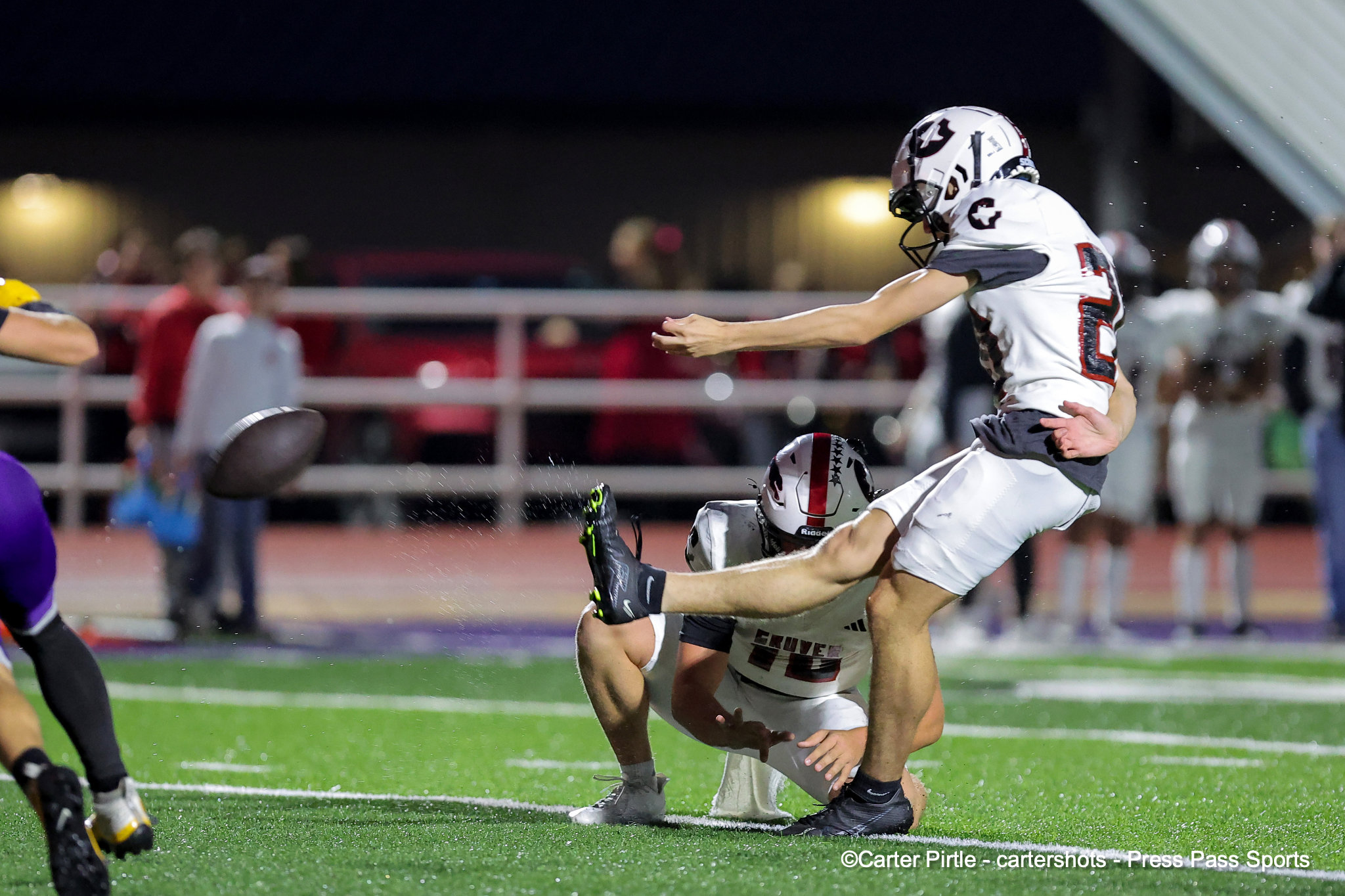 Football players compete for the ball during a night game on a bright green turf; one player in white jumps forward.