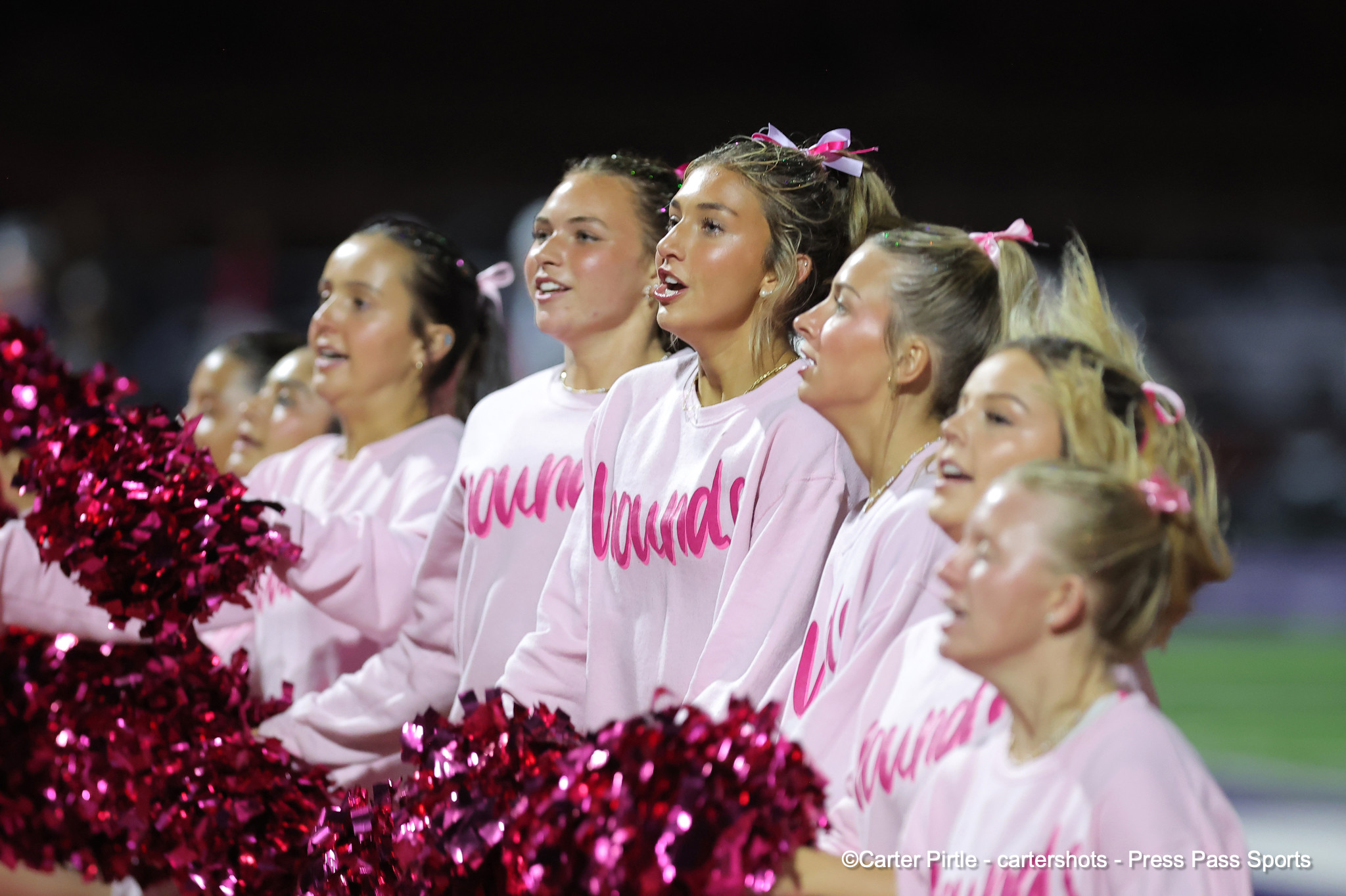 Group of cheerleaders in pink shirts cheering with pink pom-poms on a field at night.