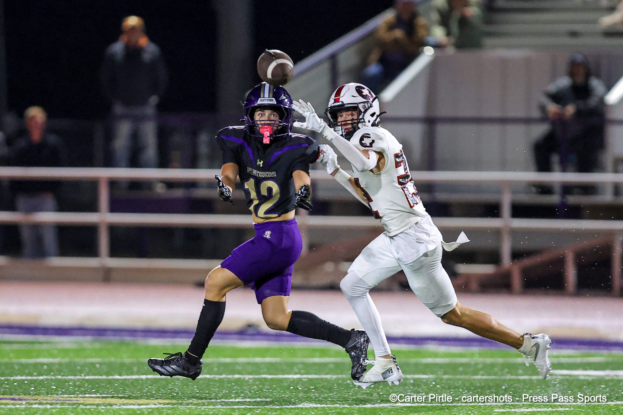Two football players sprint for a rising ball during a night game on green turf.