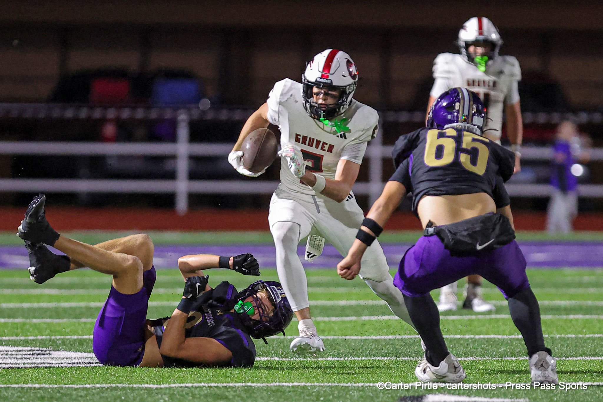 Football player in a white jersey carries the ball while being pursued by a purple-clad defender on a green turf field at night.
