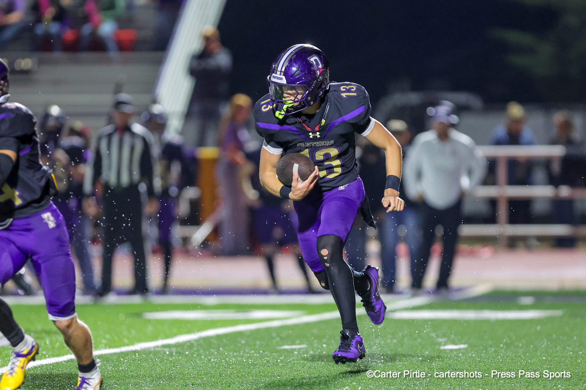 Football player in purple uniform sprinting with the ball on a bright turf field at night fall game.