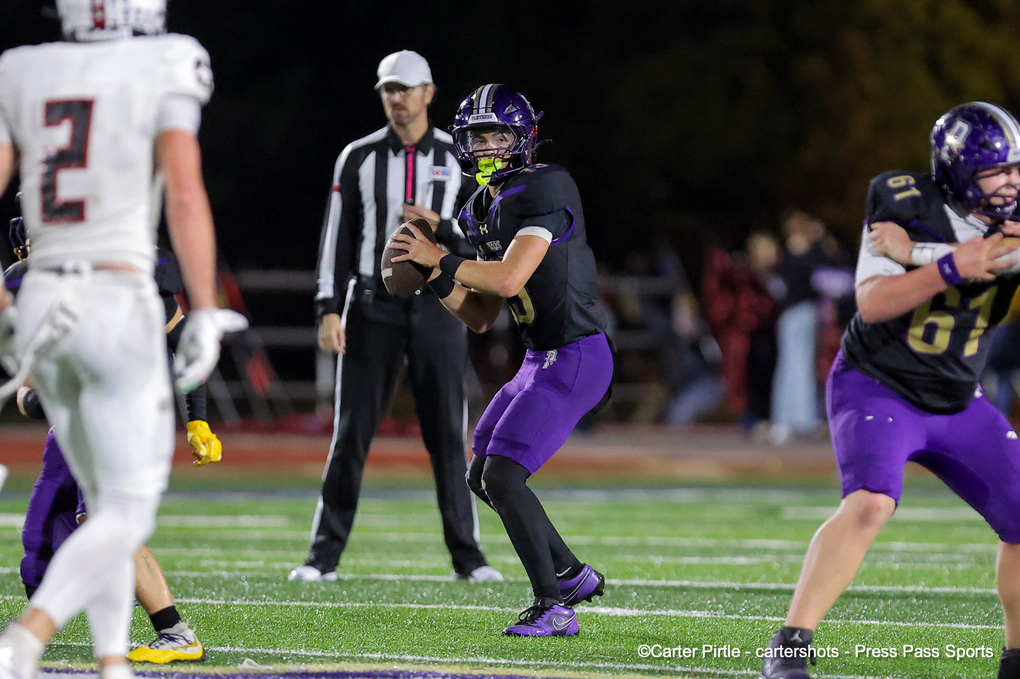 Quarterback in purple and black uniform prepares to throw a pass during a night football game with a referee and other players nearby.