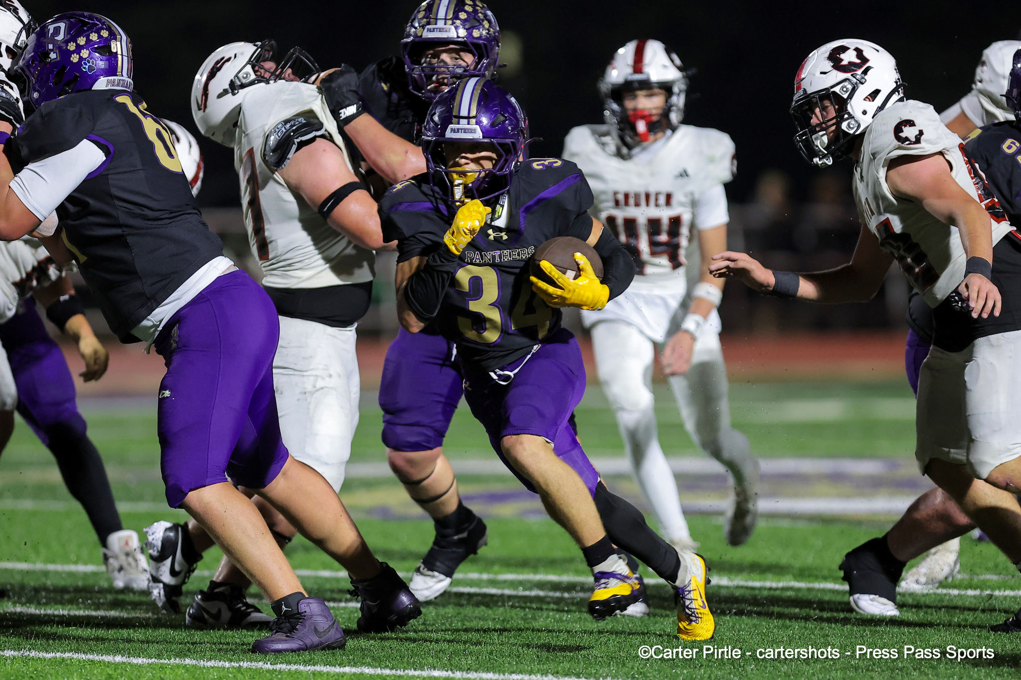 A football running back in purple and yellow sprints with the ball as defenders close in on a night game on a green field.