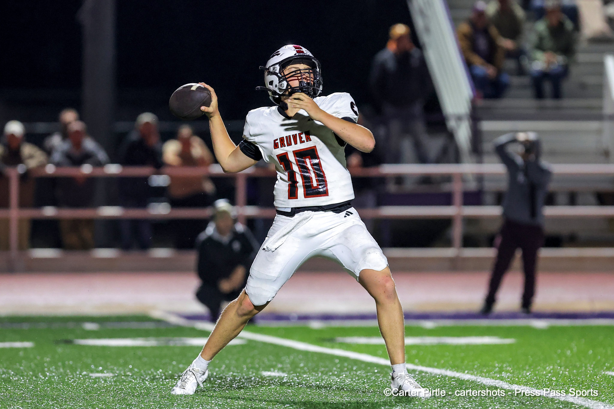 Quarterback in white jersey number 10 throws a pass on a bright football field at night, with blurred spectators in the background.