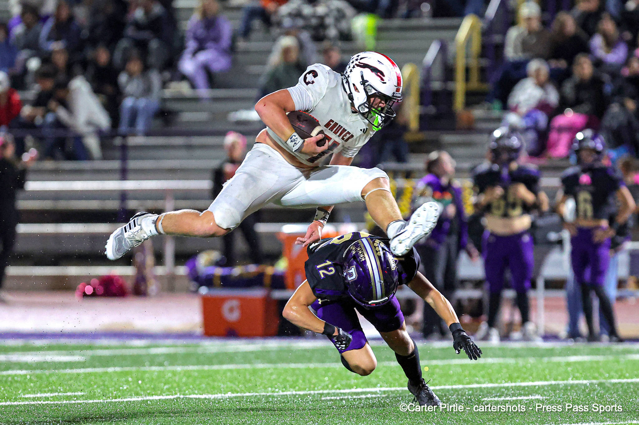 White-jersey football player leaps with ball, dodging a purple-clad defender on a bright turf field at night.