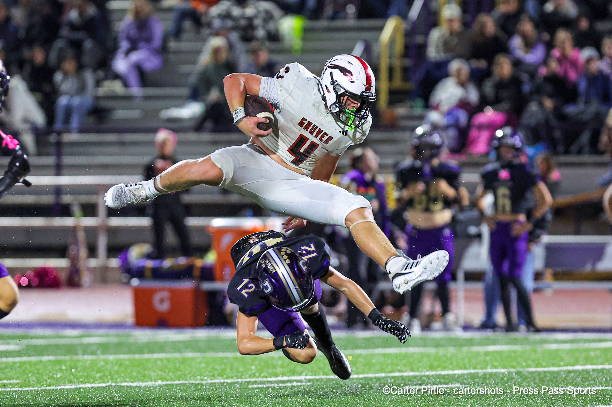White-jersey football player wearing number 4 leaps over a purple-clad defender with the ball during a night game. Crowd blurred in the stands behind.
