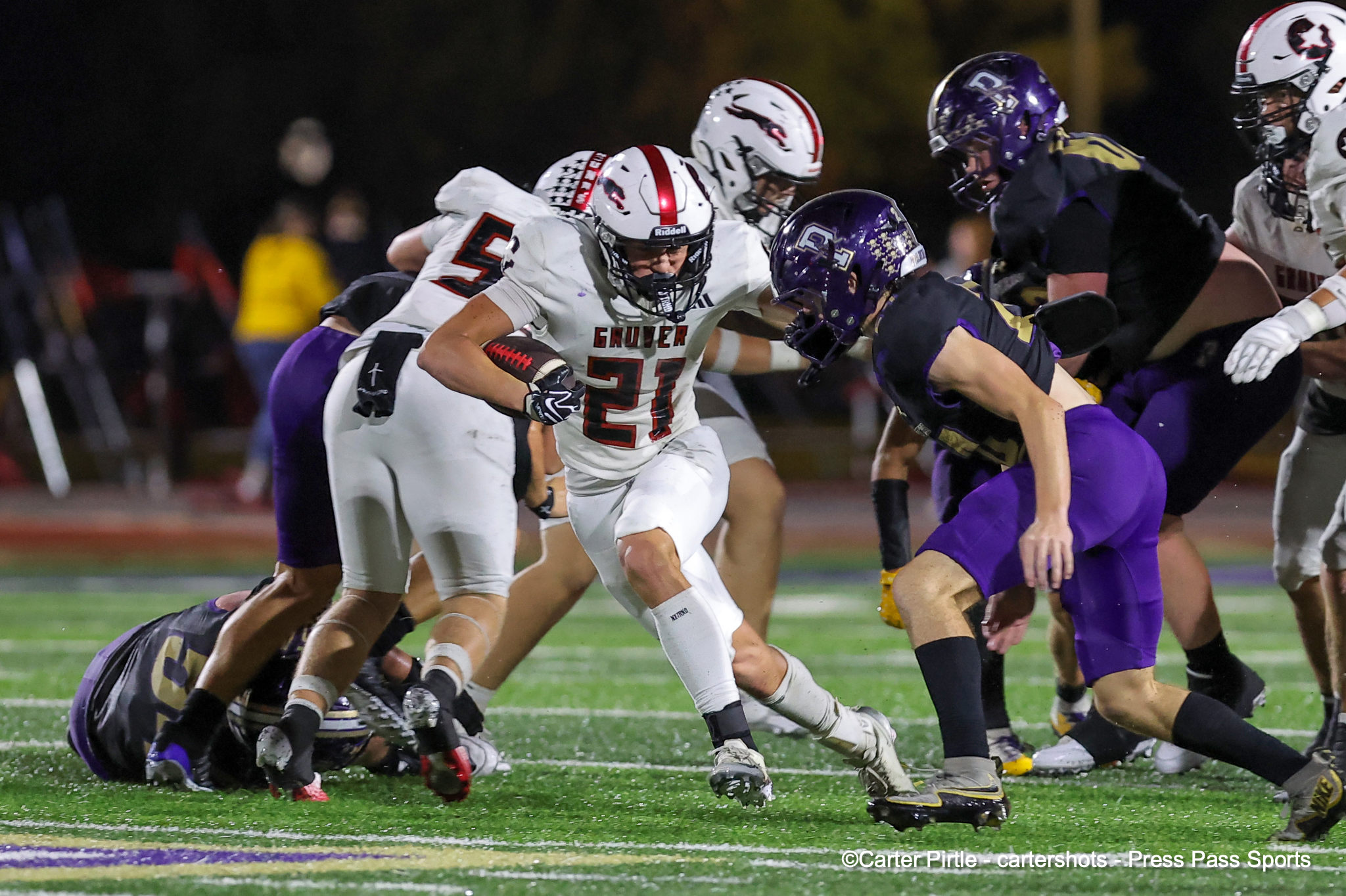 Players in white and purple tackle each other during a night football game on a turf field.