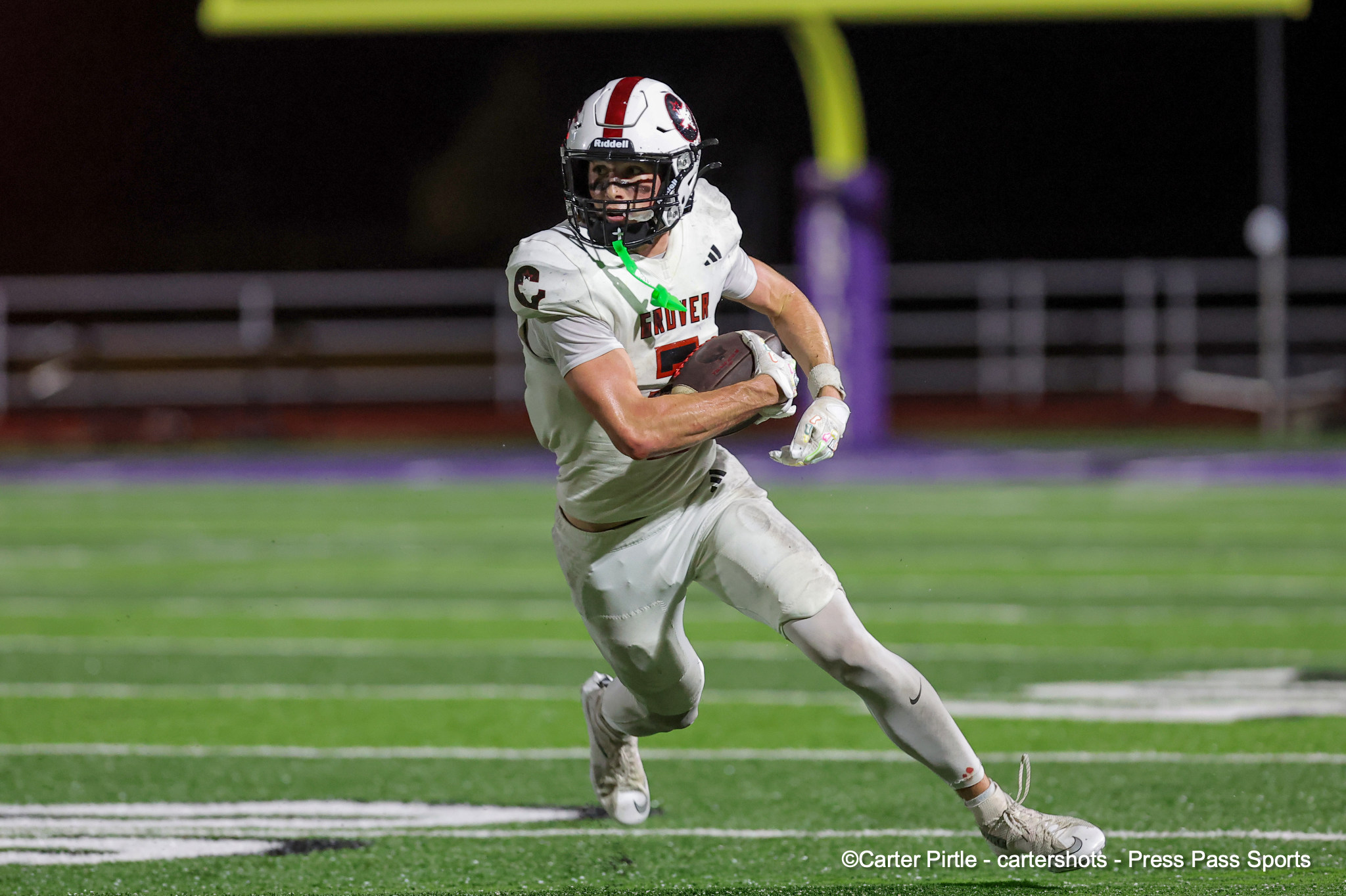 Football player in white uniform sprinting with ball during a night game on a green turf field, neon goal post in the background