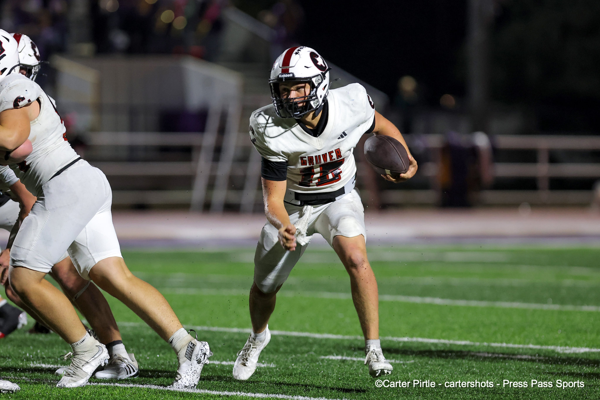 Football game action: running back in a white jersey clutches the ball and races down the field at night.
