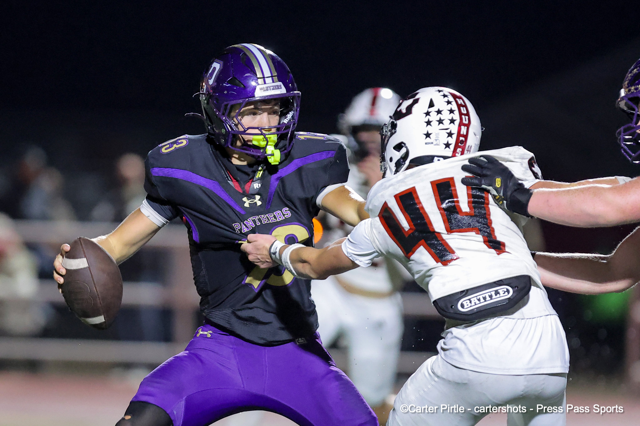 Nighttime football action: a purple Panthers runner grips the ball and charges forward while a defender in white reaches in for a tackle.